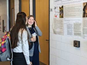 Two women stand by a research poster taped to a white wall. One woman is smiling; she touches her face with her left hand and holds a cup of coffee with her right hand. The face of the woman she is speaking with is not seen; only the back of her head is visible.