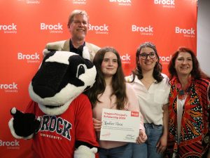 Two adults and two high school students pose for a photo in front of a branded banner with a university mascot.