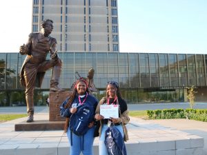 Two high school students pose for a photo in front of a bronze statue with a grey building in the background.