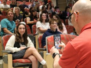 A father takes a photo of his teenage daughter using a cellphone camera in an auditorium.