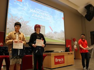 Two high school students hold award certificates and pose for photo at the front of an auditorium.