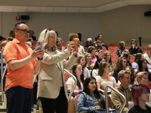 Two adults take photos using their cell phones from the audience seating in an auditorium.