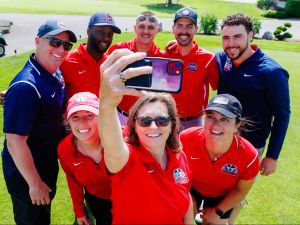 A group of eight individuals take a group selfie photo outdoors on a golf course.