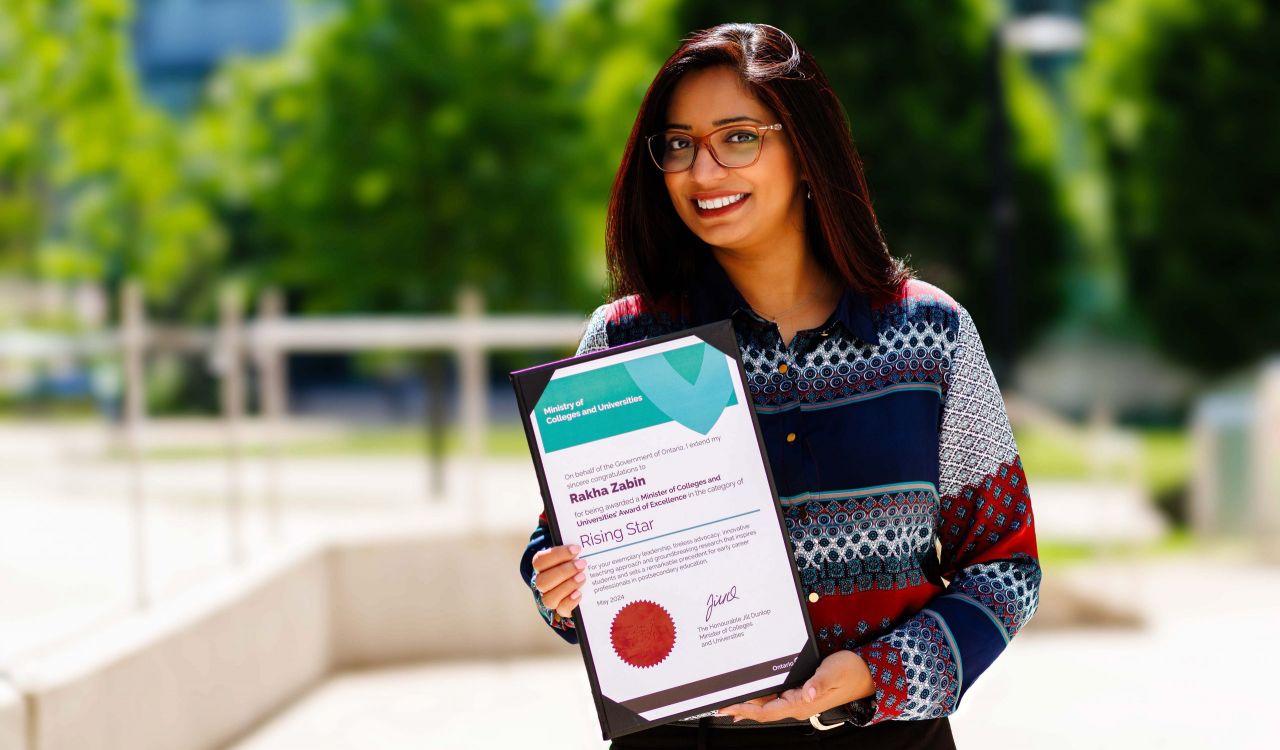 A woman stands outside at Brock’s main campus on a sunny day with blurred trees in the background. Shown from the waist up, she is smiling warmly while holding an award certificate in her hands.