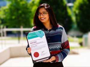 A woman stands outside at Brock’s main campus on a sunny day with blurred trees in the background. Shown from the waist up, she is smiling warmly while holding an award certificate in her hands.