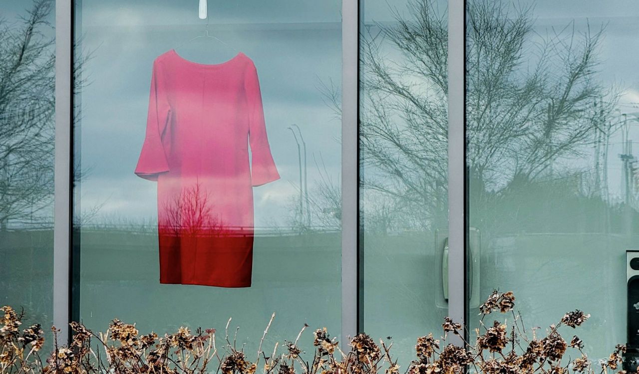 A view from outdoors shows an empty red dress hanging in the window of the Marilyn I. Walker School of Fine and Performing Arts at Brock.