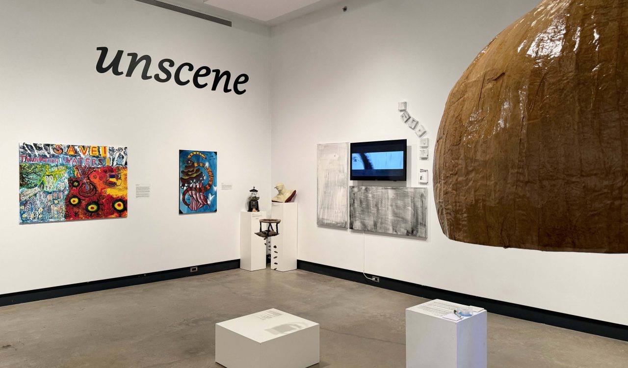 Colourful paintings, a large sculpture of an onion painted brown, along with a video screen and black and white paintings adorn the walls of an art gallery. A large title in black lettering showing the word unscene is on the wall of the gallery.