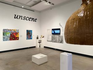 Colourful paintings, a large sculpture of an onion painted brown, along with a video screen and black and white paintings adorn the walls of an art gallery. A large title in black lettering showing the word unscene is on the wall of the gallery.