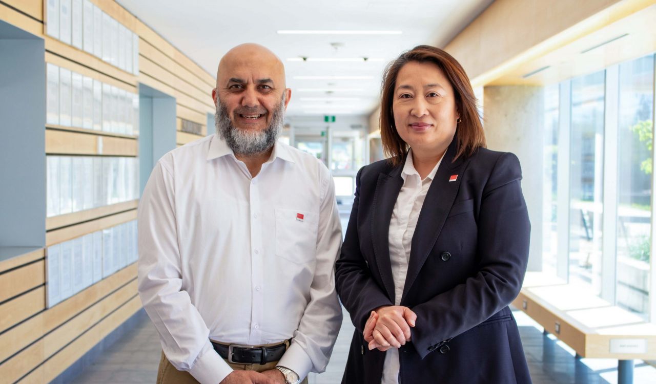 A man and a woman dressed in business clothes stand smiling at the camera in a hallway with frames on the wall on one side and windows on the other.