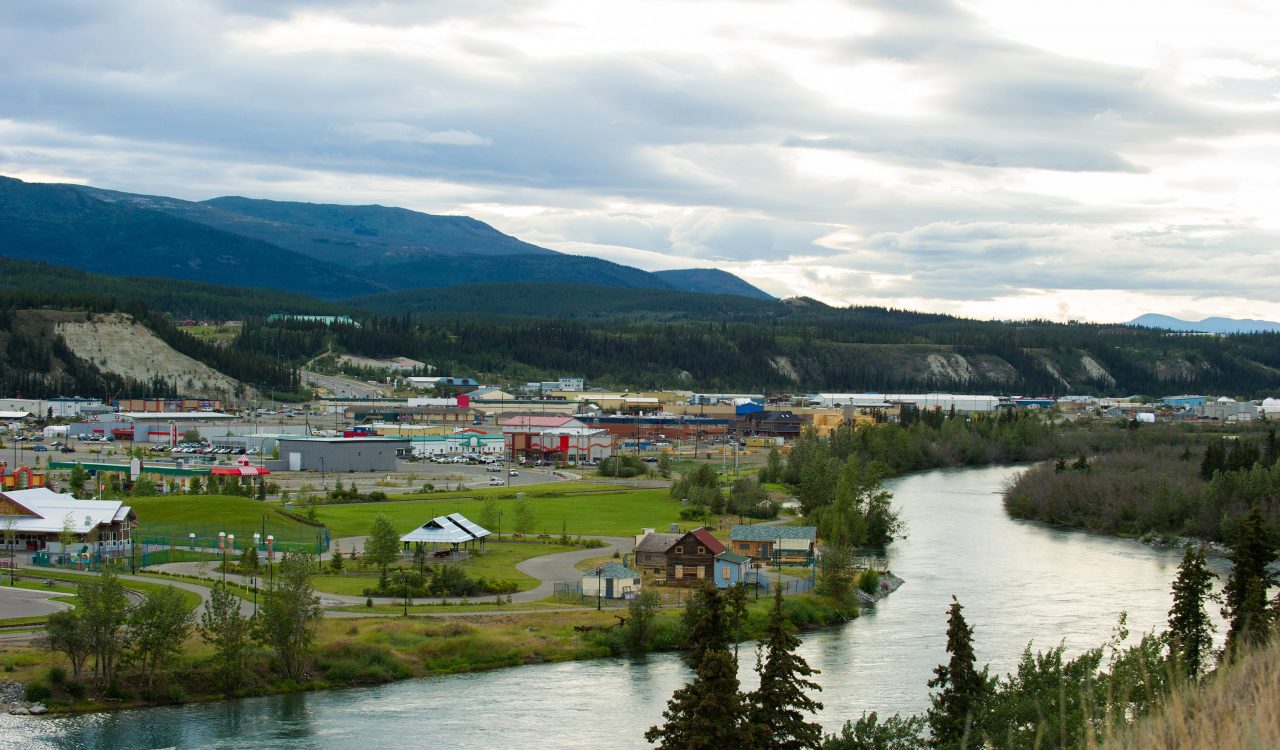 Landscape of Whitehorse, Yukon, Canada. A narrow river curves alongside a small town. Mountains line the landscape in the background.