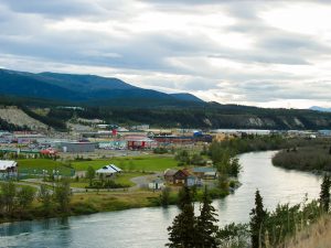 Landscape of Whitehorse, Yukon, Canada. A narrow river curves alongside a small town. Mountains line the landscape in the background.