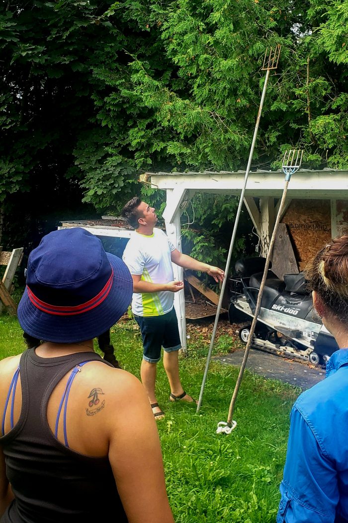 A man stands next to two large fishing spears leaning against a small building. A small group of people are listening to him talk about the spears.