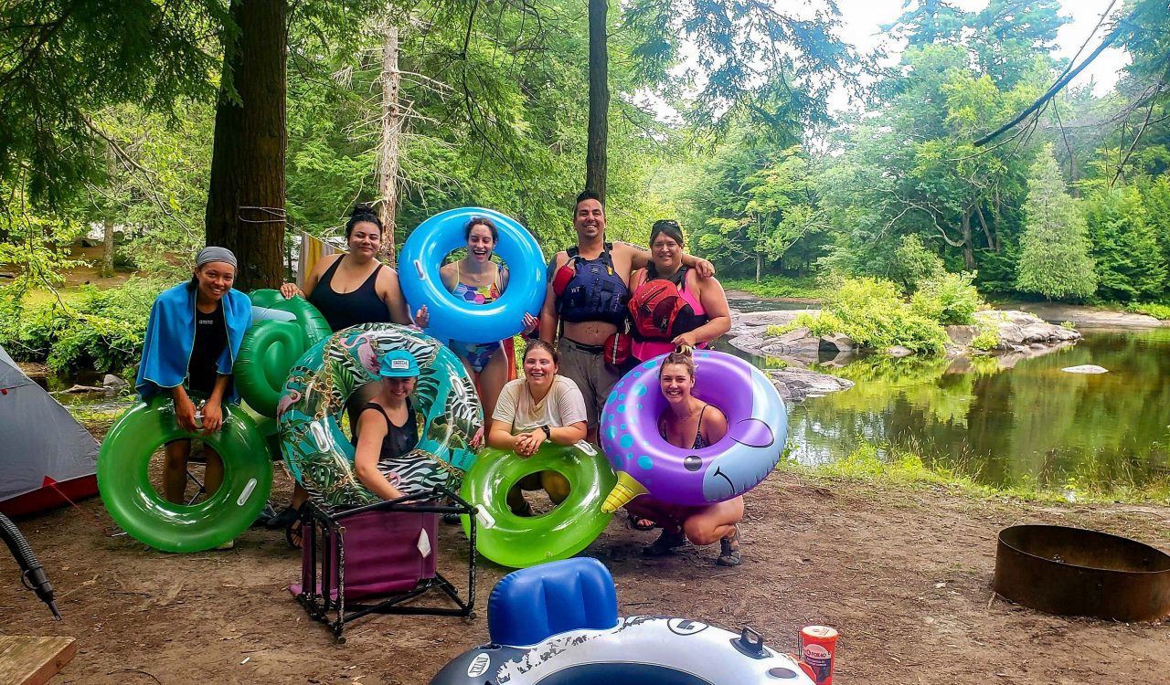 Eight people pose for a photo with colourful inner tubes outside next to a tree-lined river.