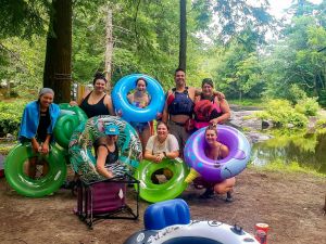 Eight people pose for a photo with colourful inner tubes outside next to a tree-lined river.