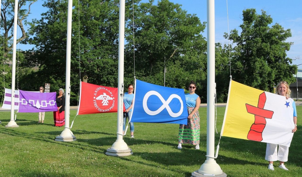 Five women stand in a row on a university campus, each woman is holding up one flag.