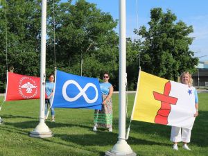 Five women stand in a row on a university campus, each woman is holding up one flag.