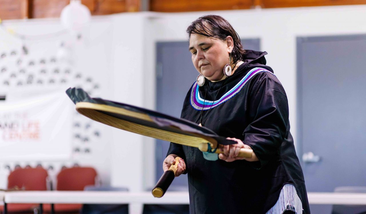 An Inuk woman dressed in traditional regalia drums on a traditional Inuit percussion instrument made with a wooden frame, protruding handle and a synthetic membrane stretched across the frame.