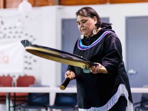 An Inuk woman dressed in traditional regalia drums on a traditional Inuit percussion instrument made with a wooden frame, protruding handle and a synthetic membrane stretched across the frame.