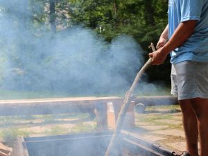 A young First Nations man tends to a fire in a pit with a large stick.