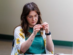 A woman sits at a table holding a metal ring that she is weaving with sinew into a pattern that resembles a spider’s web.