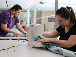 A woman stands over a piece of fabric holding a measuring tape while a seated woman sews ribbons onto a skirt using a singer sewing machine.