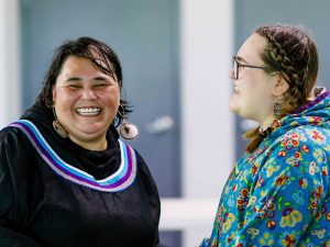 A mother and teenage daughter dressed in traditional Inuit regalia face each other laughing.