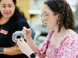 A woman sews a seal fur bracelet while another woman, blurred in the background, looks on.