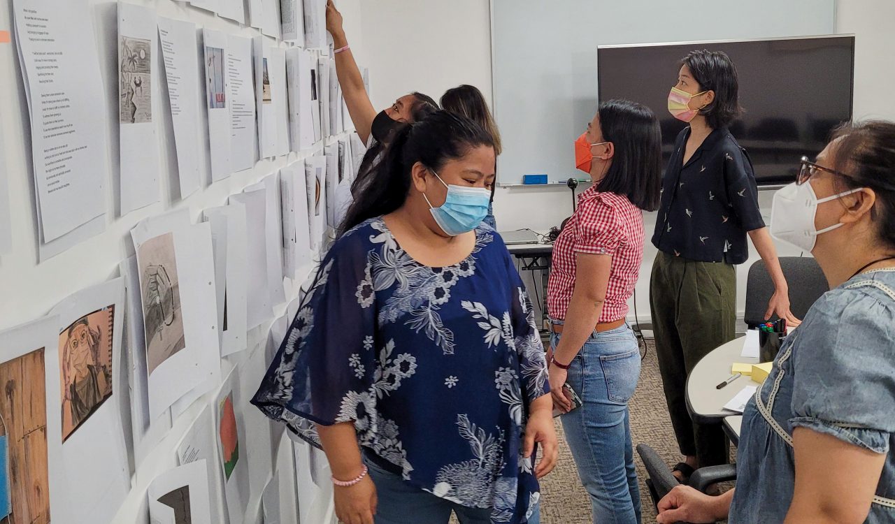 The creative team behind Ingat: An Anthology of Works by Migrant Domestic Creatives in Hong Kong studies a wall full of separate pieces during a meeting at the University of Hong Kong. Each person is masked in the conference room.