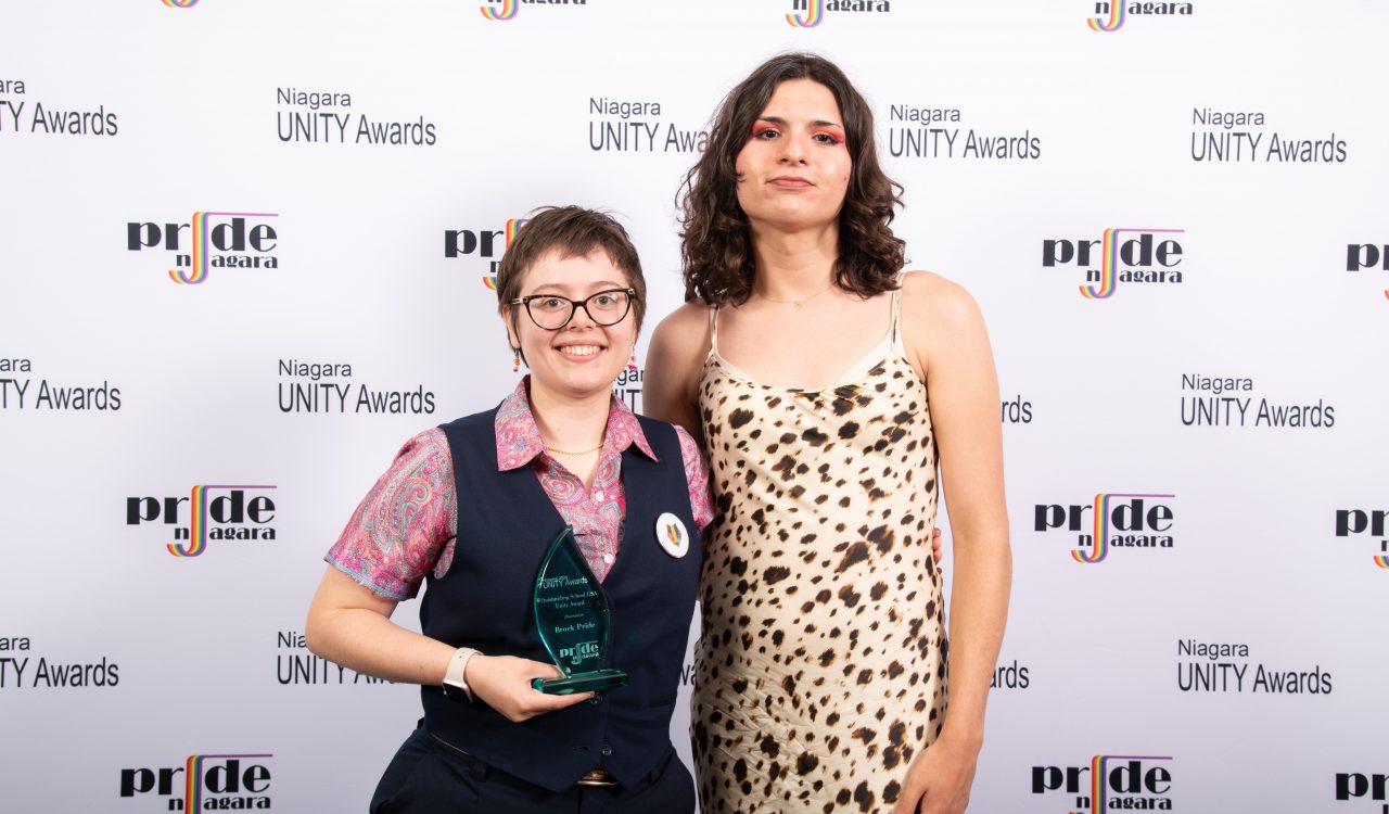 Two people pose for a photo with an award in front of a logo backdrop.