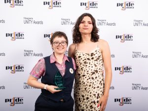 Two people pose for a photo with an award in front of a logo backdrop.