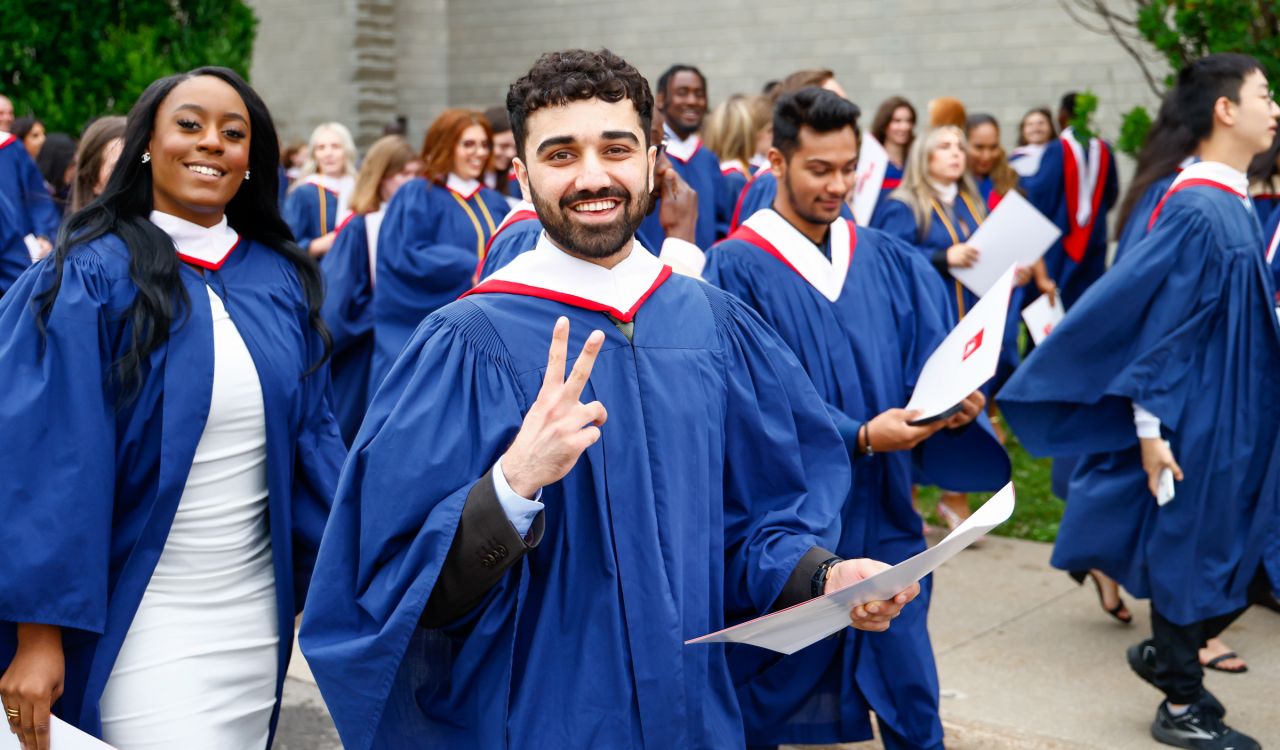 A crowd of people in graduation gowns walk outside - on flashing a peace sign.