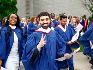 A crowd of people in graduation gowns walk outside - on flashing a peace sign.