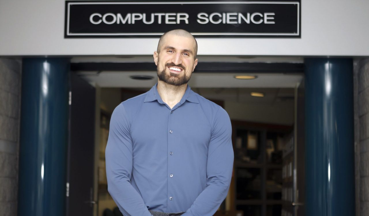 Brock University Computer Science professor Ali Emami stands in a hallway in front of a sign that reads “Computer Science.”