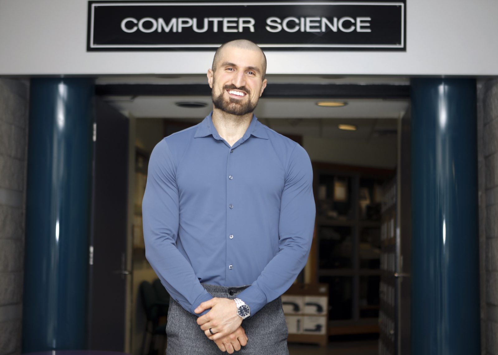 Brock University Computer Science professor Ali Emami stands in a hallway in front of a sign that reads “Computer Science.”