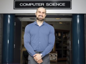 Brock University Computer Science professor Ali Emami stands in a hallway in front of a sign that reads “Computer Science.”