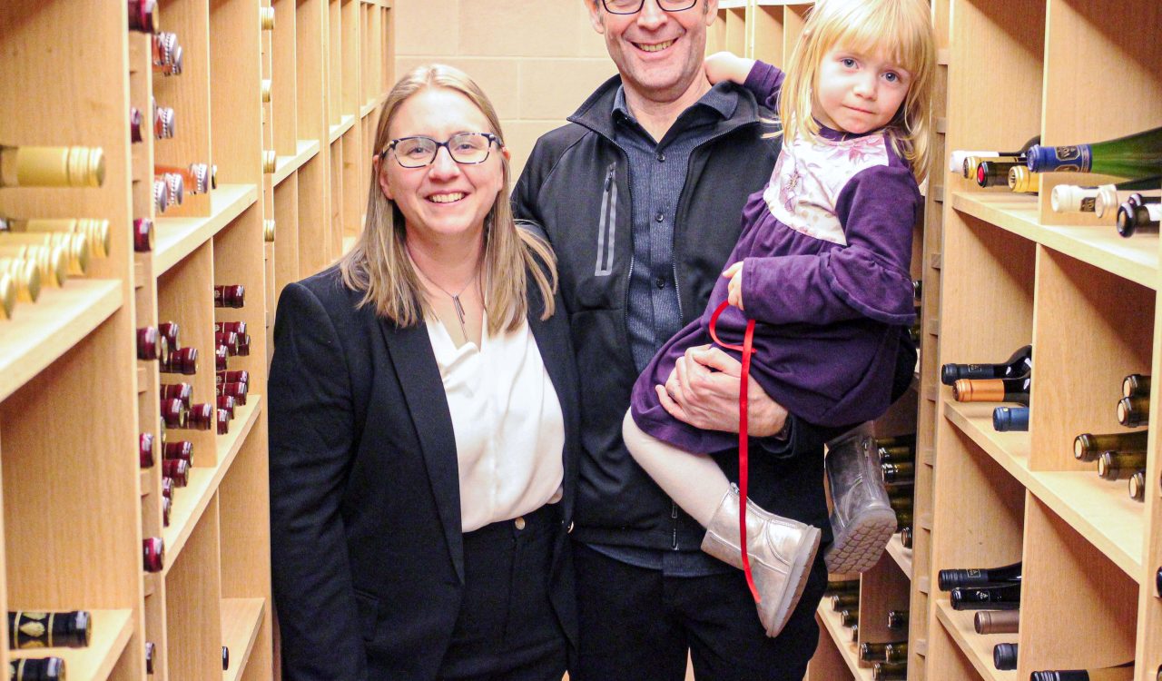 A woman, man and their daughter smile while in a wine cellar.