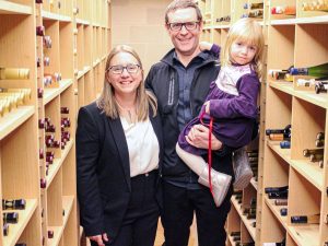 A woman, man and their daughter smile while in a wine cellar.