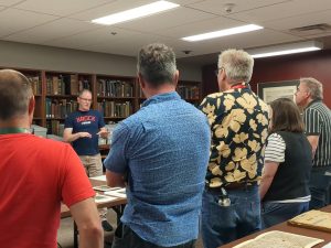 A group of six people stand in front of a row archival books listening to a man speak. 