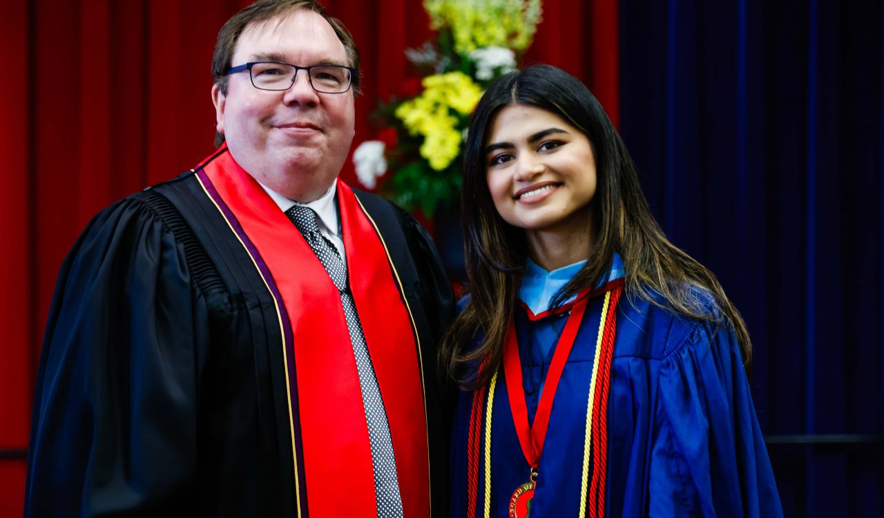 A man and a woman in academic robes pose for a photo on stage during a university graduation ceremony.