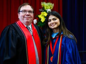 A man and a woman in academic robes pose for a photo on stage during a university graduation ceremony.