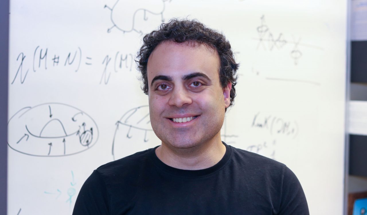 Head-and-shoulders photo of Assistant Professor of Physics Barak Shoshany smiling into the camera with an out-of-focus board of physics calculations in the background.