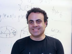 Head-and-shoulders photo of Assistant Professor of Physics Barak Shoshany smiling into the camera with an out-of-focus board of physics calculations in the background.