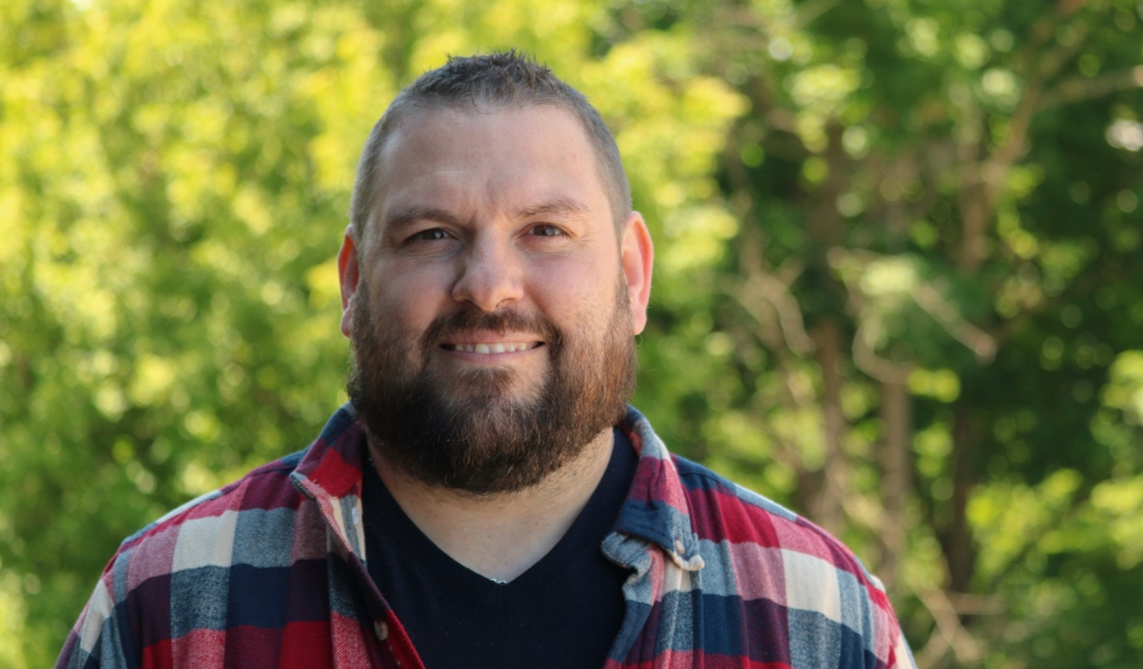 A man in a plaid shirt stands in front trees.