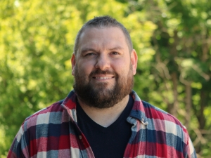 A man in a plaid shirt stands in front trees.