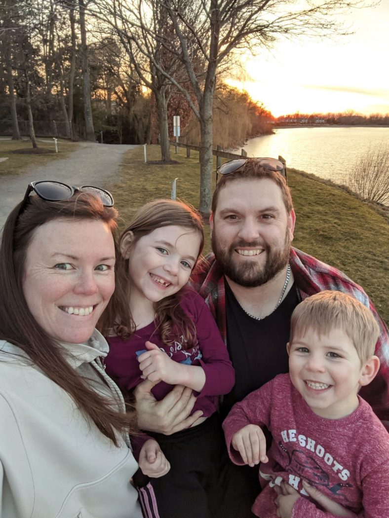 A husband and wife pose with their two children in a park near a lake.