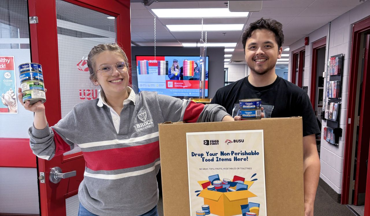 Two university students hold up a box and non-perishable food items for a photo.