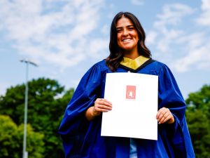 A young woman in a blue academic gown smiles for a photo outside while holding a diploma.