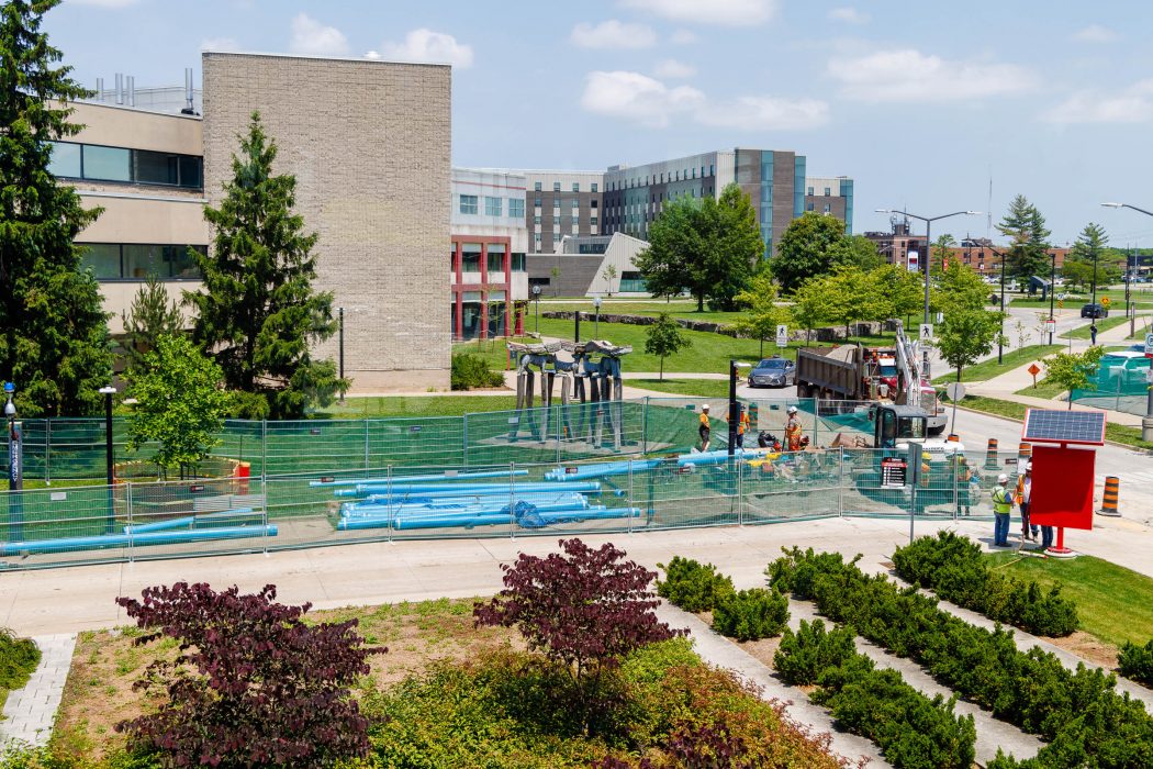 Construction workers, fences and vehicles are at the centre of Brock University's beautiful green campus under blue skies.