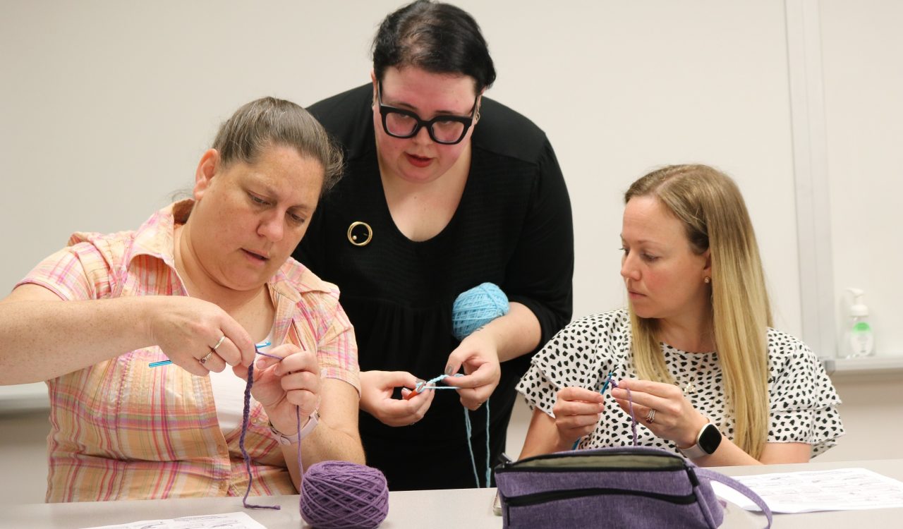 A woman stands while teaching two other women who are seated how to crochet.
