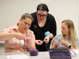 A woman stands while teaching two other women who are seated how to crochet.